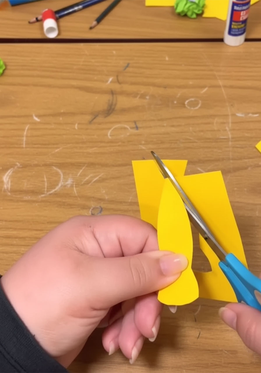 Child cutting a yellow paper tie shape with scissors for the handprint Mini-Me Dad Father's Day keepsake, Step 5A, with a finished striped tie shown in foreground