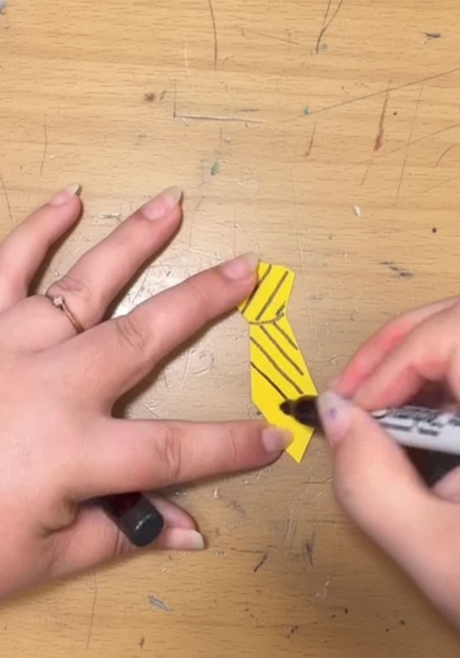 Child cutting a yellow paper tie shape with scissors for the handprint Mini-Me Dad Father's Day keepsake, Step 5B, with a finished striped tie shown in foreground