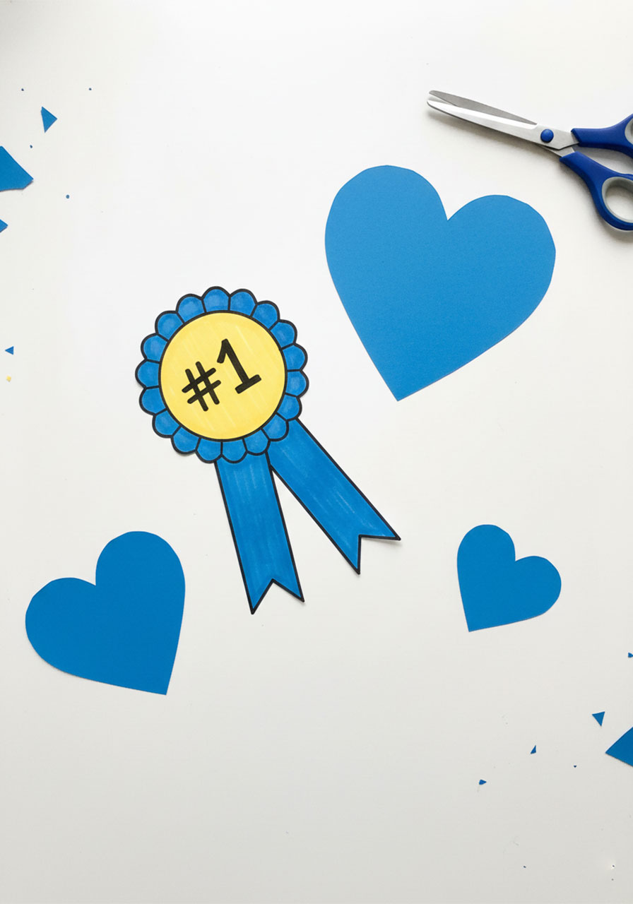 Overhead flat lay of handmade paper decorations for Father's Day card including blue and yellow number 1 award ribbon and three blue paper hearts