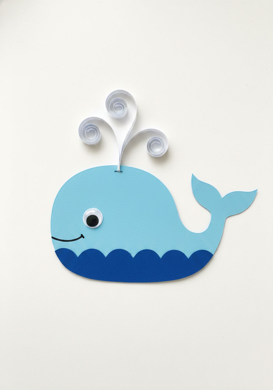 Fully assembled light blue paper whale for Father's Day pop up card showing googly eye, hand-drawn smile, curled white waterspout strips, and dark blue scallop wave base