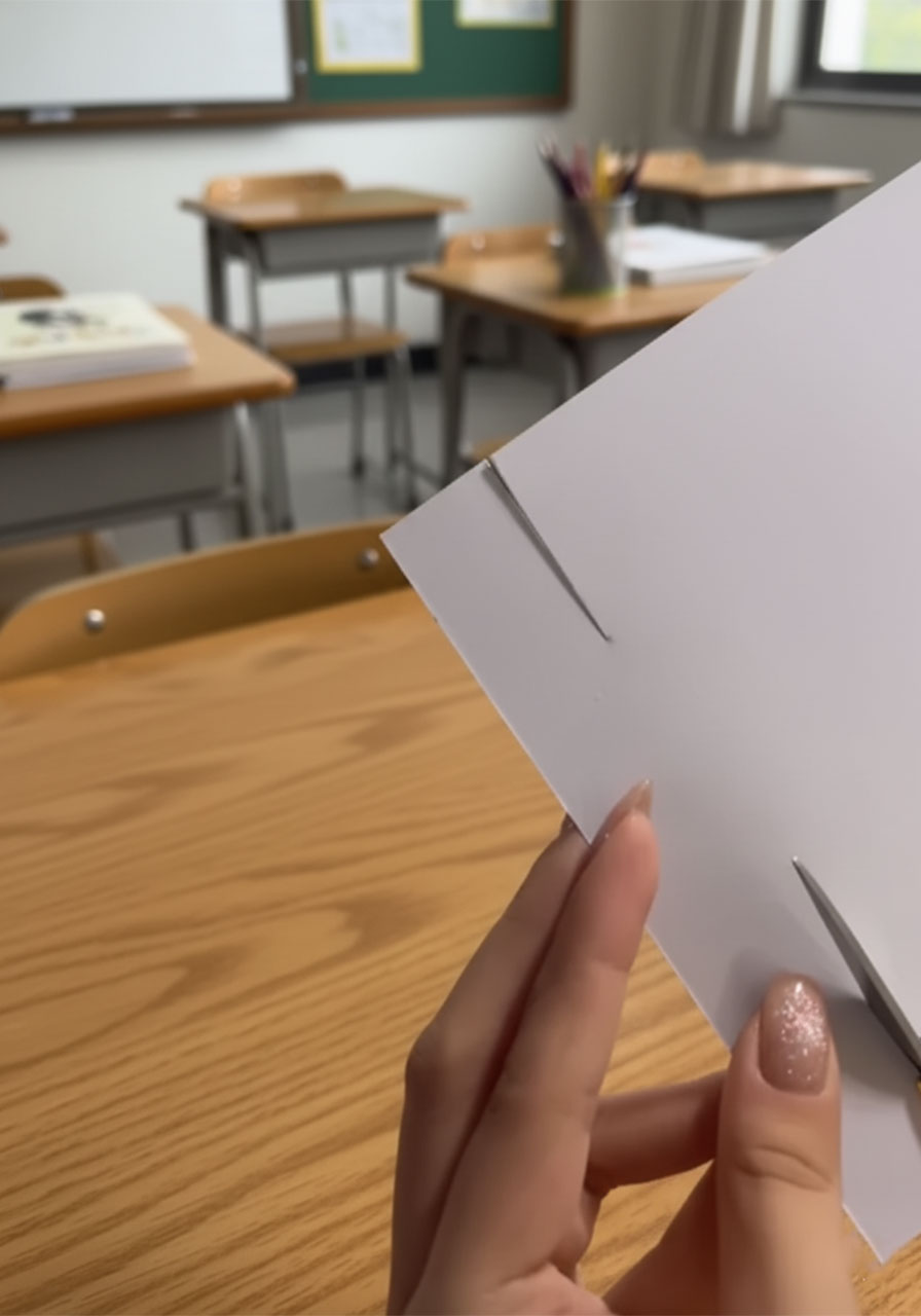Hands folding white paper into a V-shaped shirt collar for a DIY Father's Day card insert, showing the diagonal crease mid-fold step2c