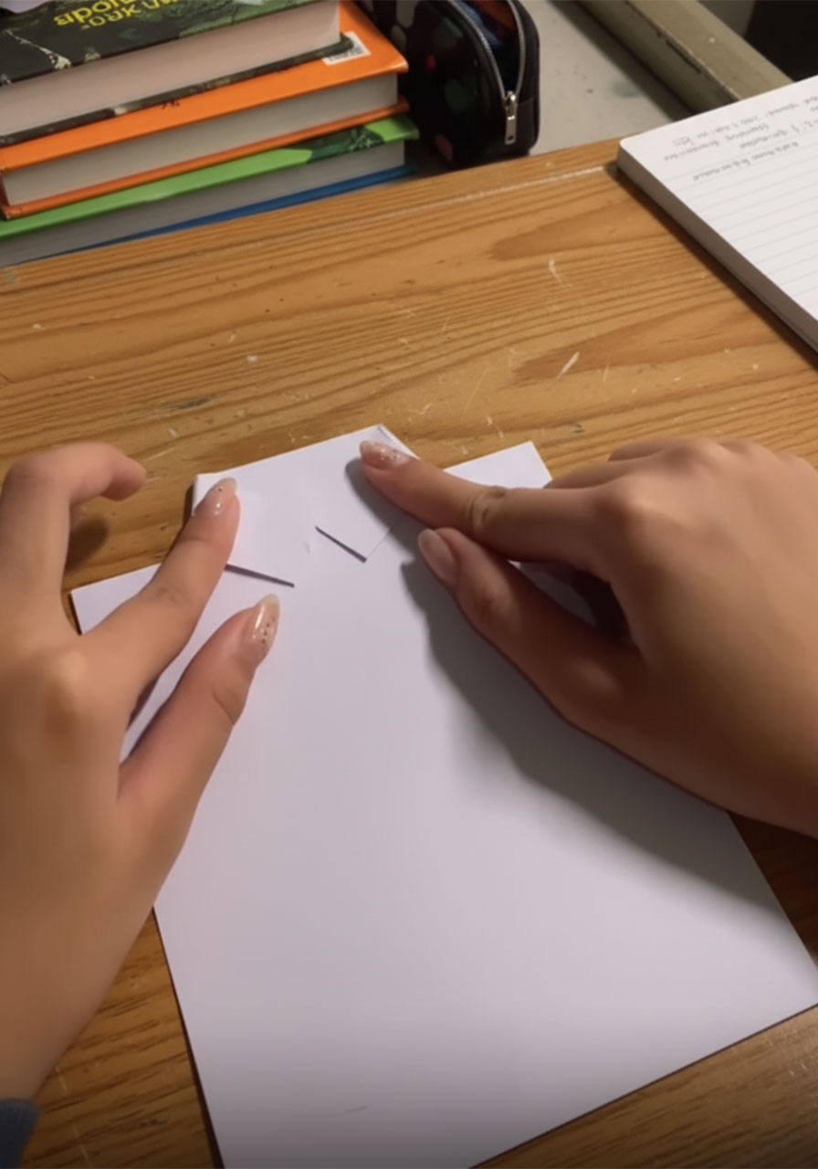 Hands folding white paper into a V-shaped shirt collar for a DIY Father's Day card insert, showing the diagonal crease mid-fold step2e