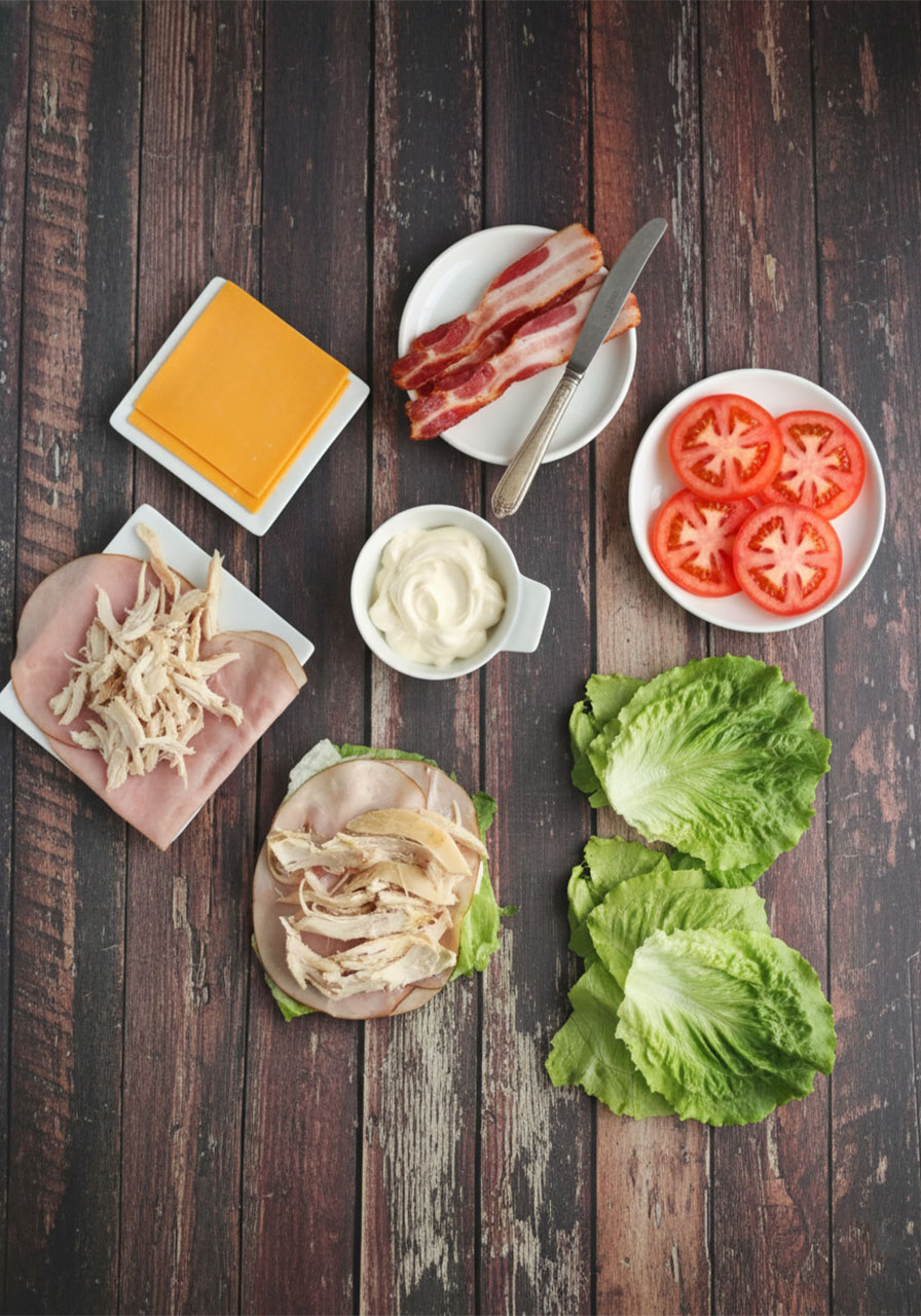 All ingredients for a keto low carb chicken club sandwich laid out flat including lettuce, chicken, bacon, ham, cheddar cheese, tomato, and mayo