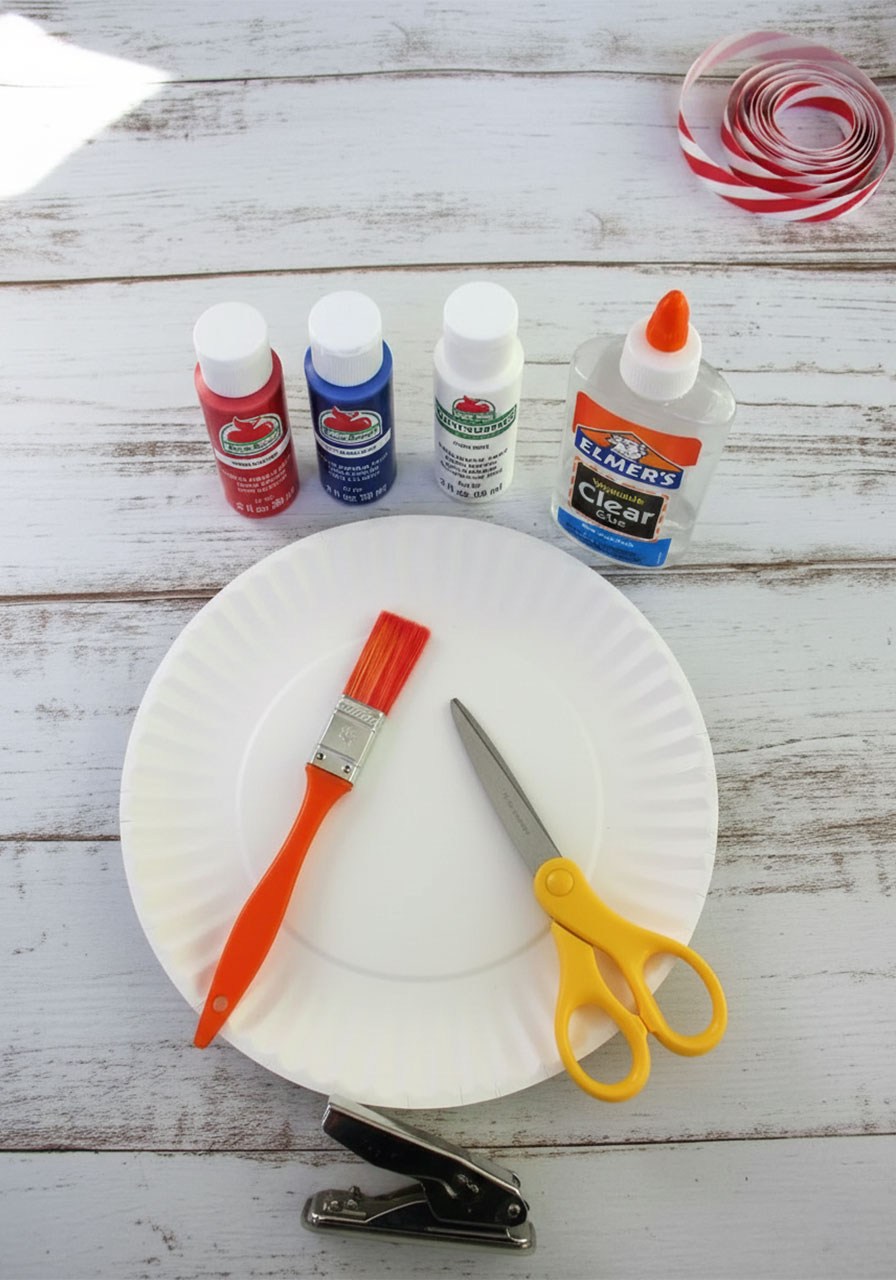 Supplies needed for patriotic paper plate wind catcher craft including paint, scissors, hole punch, and ribbon