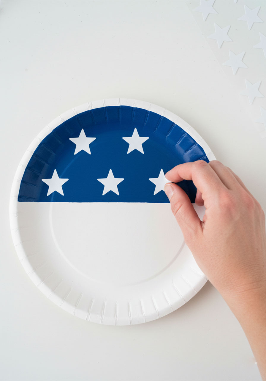 Adding white star stickers to a blue painted paper plate as step two of a patriotic paper plate flag craft for kids