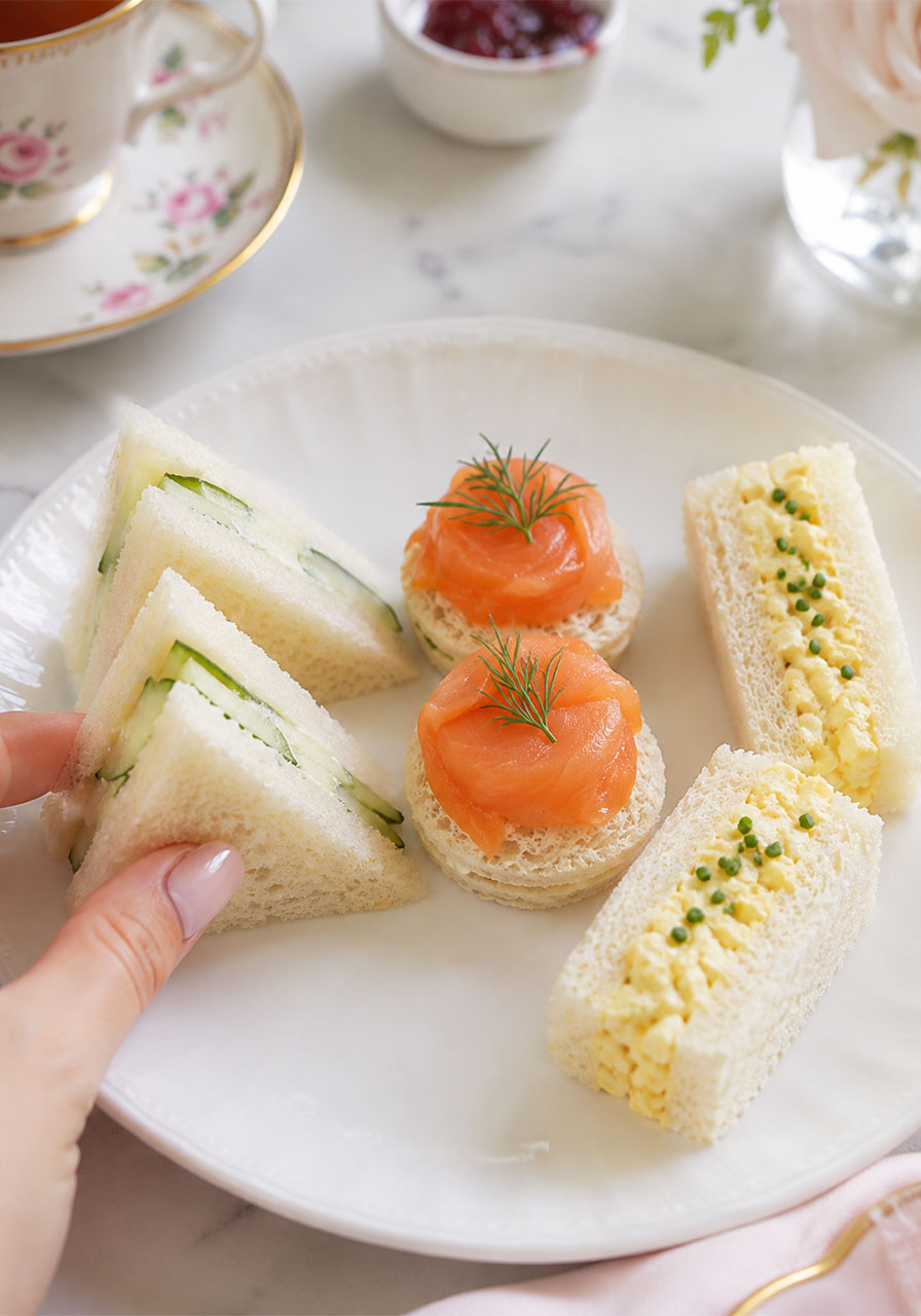 Close-up of an afternoon tea first course plate showing crustless cucumber, smoked salmon, and egg salad finger sandwiches on white china