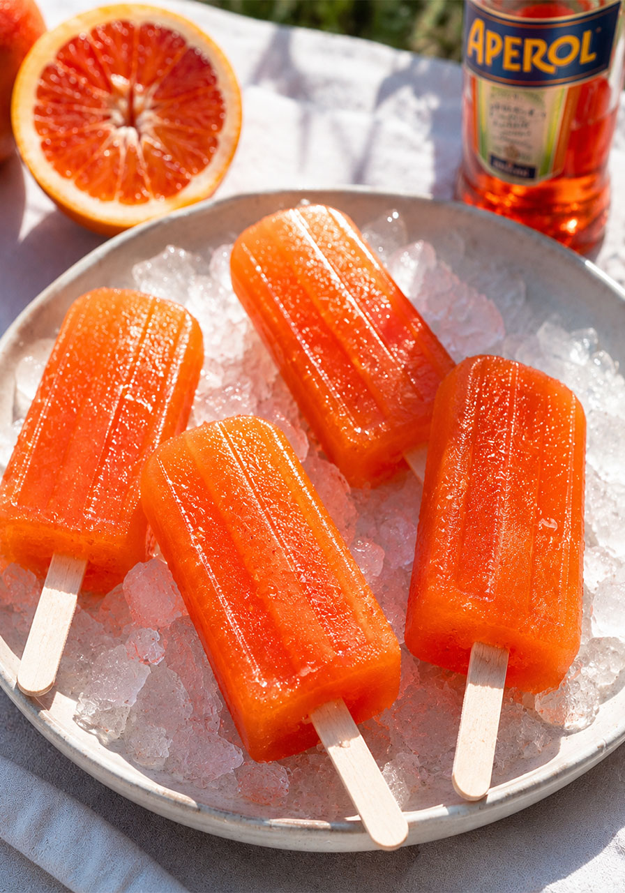 Glowing orange Aperol Spritz ice pops sweating with condensation on a plate of crushed ice, with a blood orange half and Aperol bottle in the background