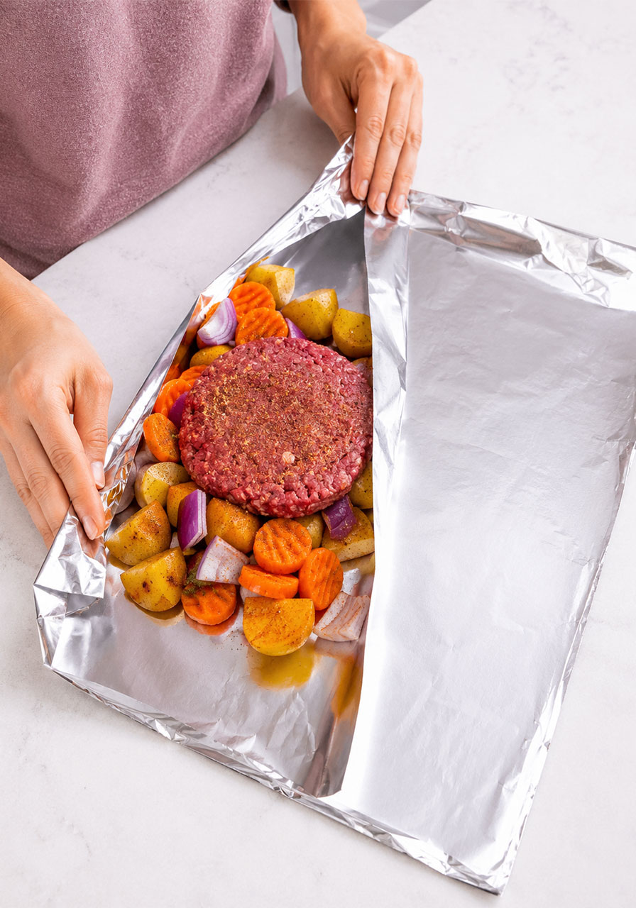 Hands assembling a campfire hobo packet by placing seasoned beef patty on top of vegetables on aluminum foil
