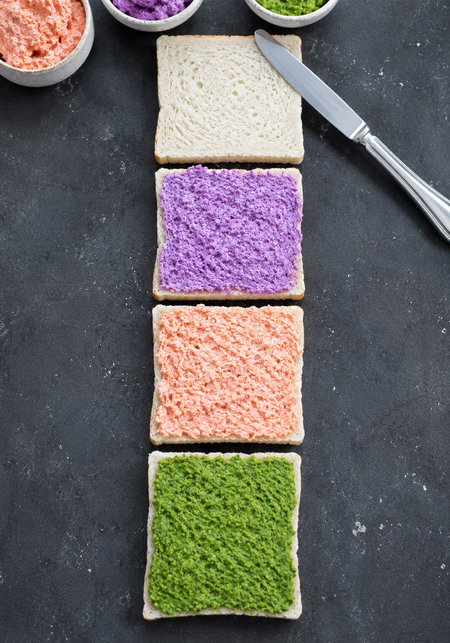 Four bread slices spread with green avocado, orange carrot, and purple cabbage yogurt layers ready to stack into tea party sandwiches