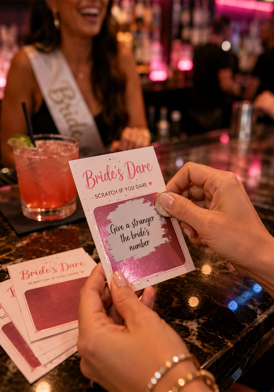Woman scratching off dare card at bar during bachelorette night out with pink cocktail and bride in background