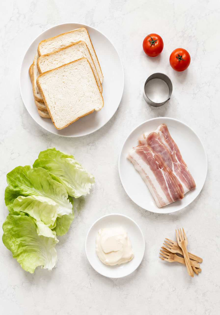 All ingredients for BLT tea sandwiches laid out flat including thin white bread, cocktail tomatoes, bacon, butter lettuce, mayo, and cocktail forks