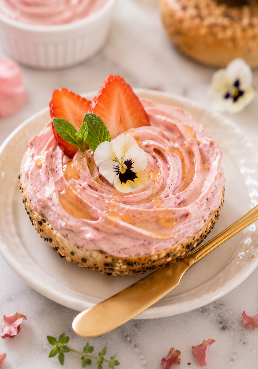 Beautifully assembled bridal shower bagel with strawberry cream cheese, honey drizzle, and edible flower on a white plate