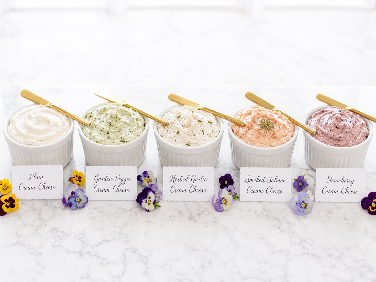 Row of labeled cream cheese ramekins with gold spreader knives for a bridal shower bagel bar