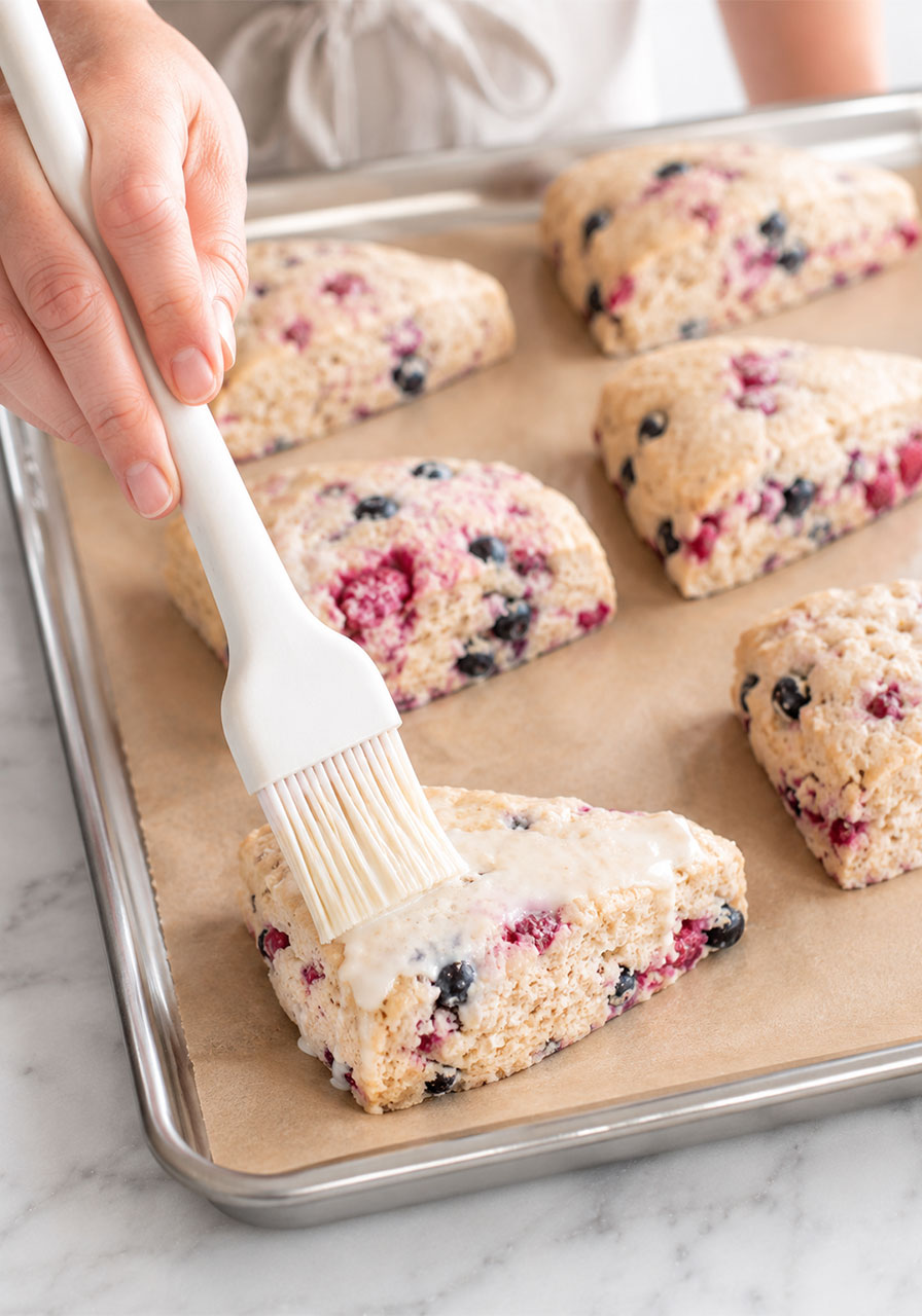 Hand brushing heavy cream onto raw mixed berry scone triangles on a parchment-lined baking sheet before baking