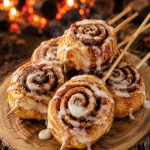 Golden campfire cinnamon roll-ups on a stick with white icing drizzle on a rustic wood slice board
