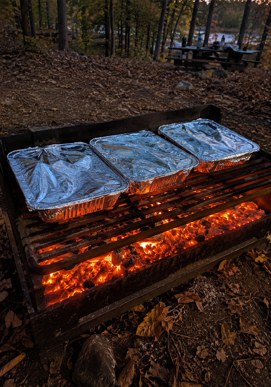 Three foil-covered nacho pans cooking on a campfire grate over glowing coals
