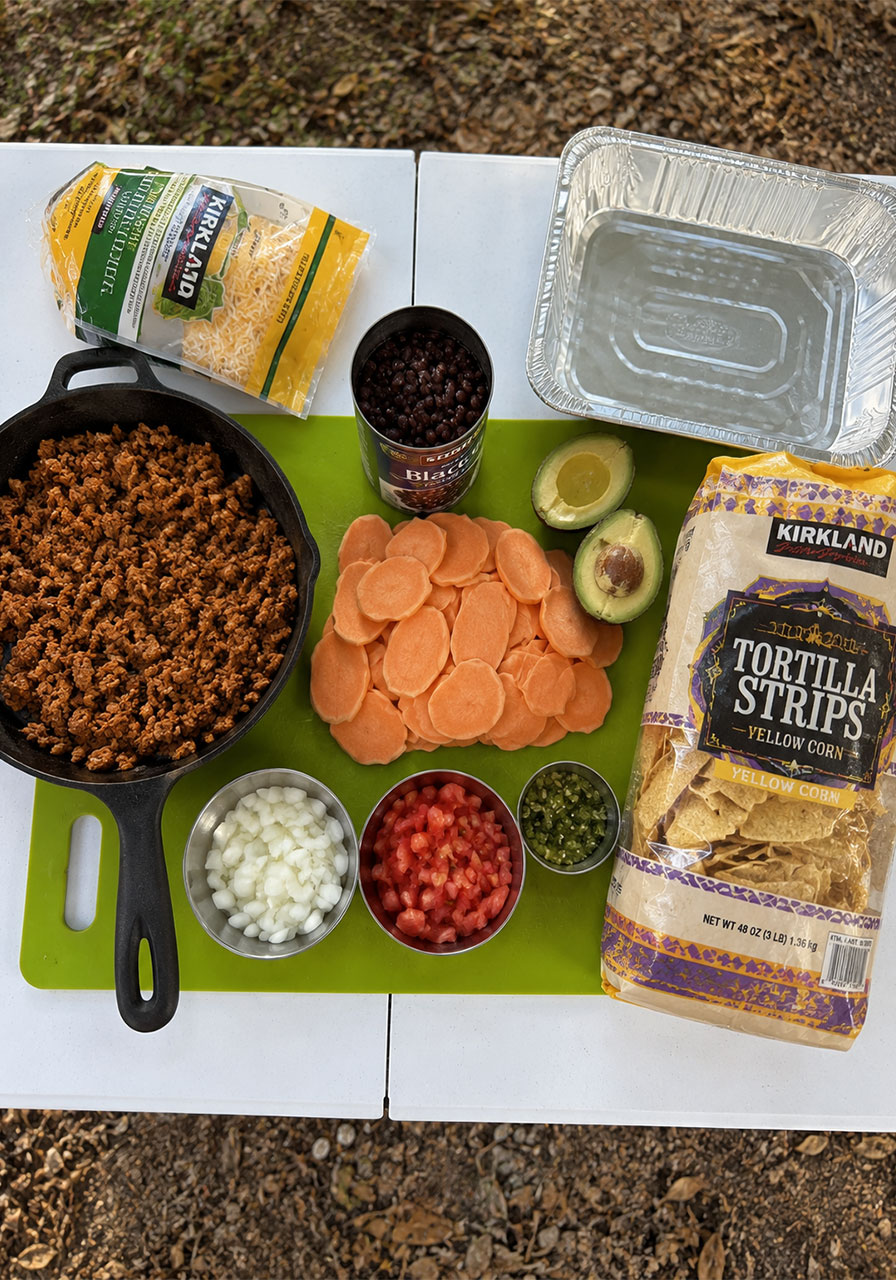 All campfire nacho ingredients prepped and laid out on a camp table including sweet potato, taco meat, black beans, avocado, and cheese