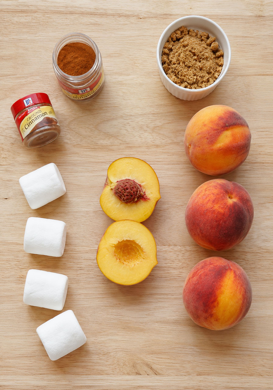 Campfire peachies ingredients: ripe peaches, large marshmallows, cinnamon, and brown sugar on a wood cutting board