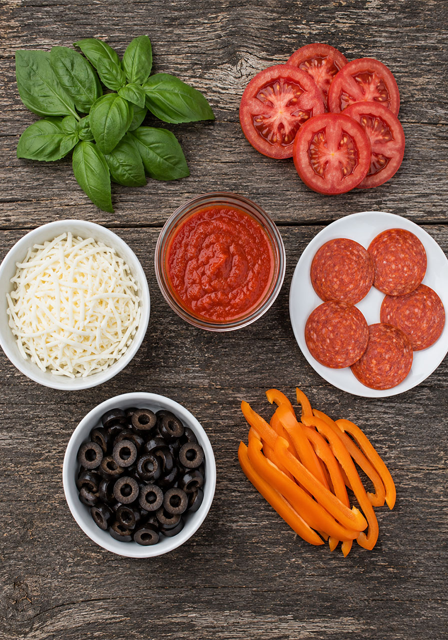 Flat lay of campfire pizza topping options including sauce, mozzarella, pepperoni, tomatoes, and basil at a campsite