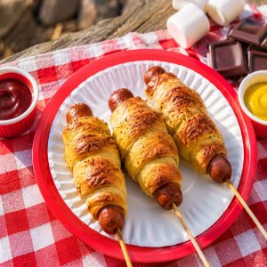 Three golden crescent-wrapped camping hot dogs on a paper plate with ketchup and mustard, cooked over a campfire