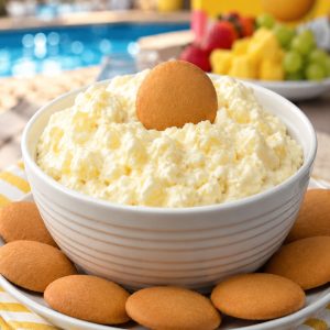 Creamy cheesecake pineapple dip in a white ribbed bowl surrounded by Nilla wafer cookies on a white plate