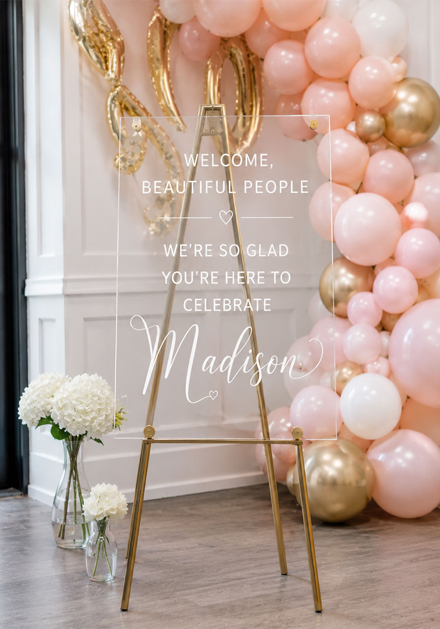 Clear acrylic welcome sign with white vinyl lettering on gold easel in front of blush balloon arch at bridal shower