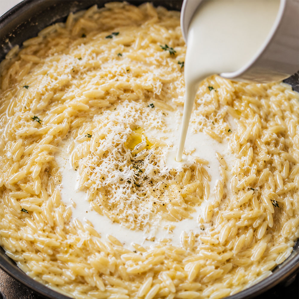 Heavy cream and parmesan cheese being stirred into cooked orzo