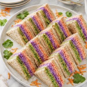 Colorful layered tea party finger sandwiches with green avocado, orange carrot, and purple cabbage yogurt spreads on a white platter