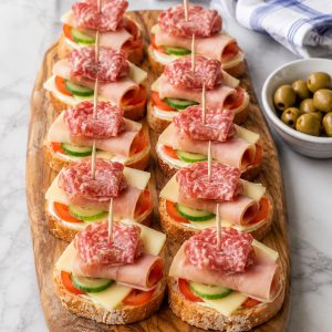 A wooden board of easy tea club sandwiches with ham, salami, cheese, tomato, and cucumber on toasted baguette slices