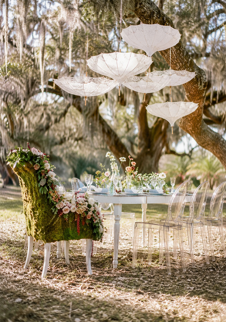 Ethereal outdoor garden bridal shower tablescape with moss-covered floral chair, ghost chairs, and parasols under oak tree