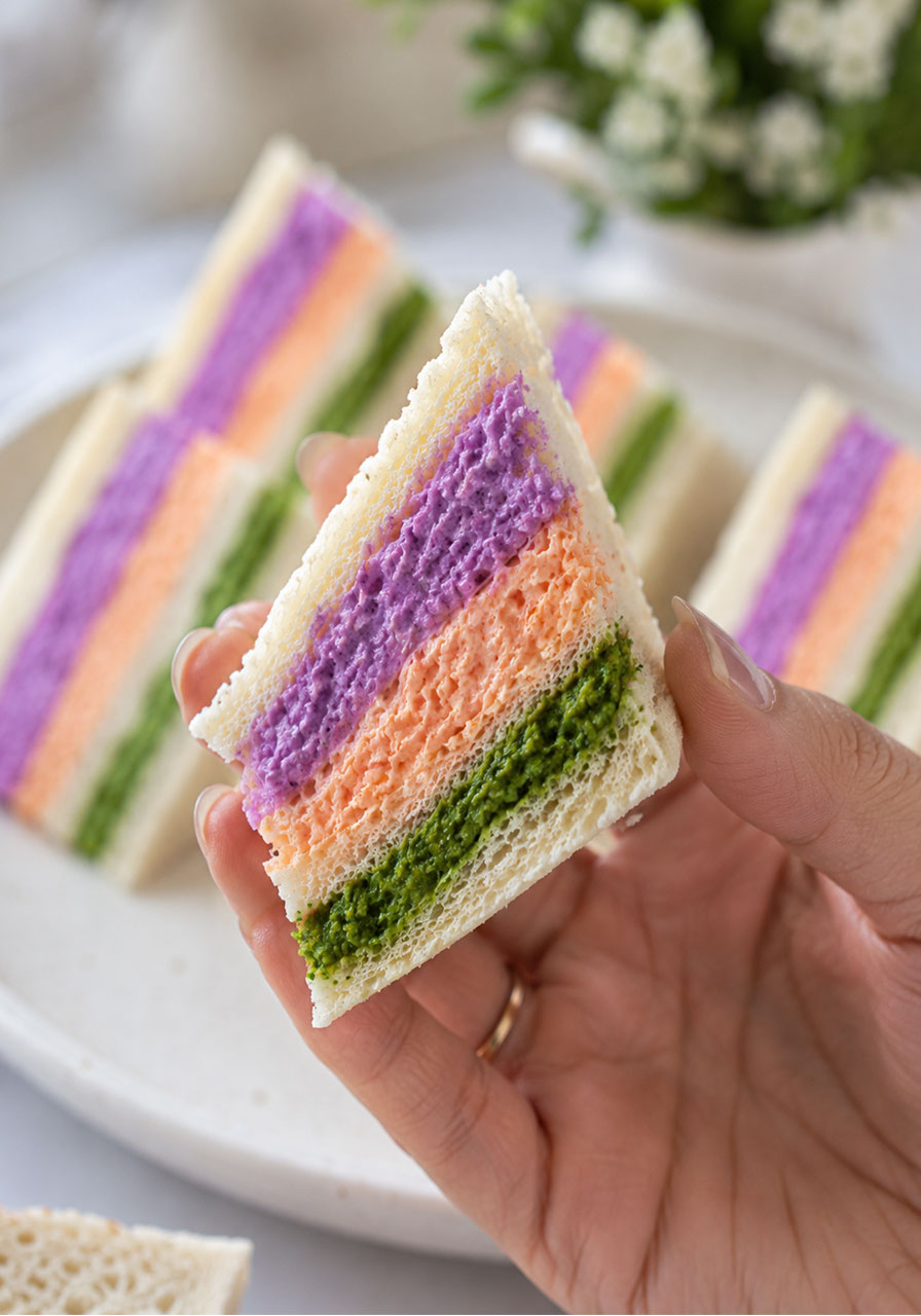 Woman holding a triangular colorful tea party finger sandwich showing bright green, orange, and purple healthy yogurt filling layers