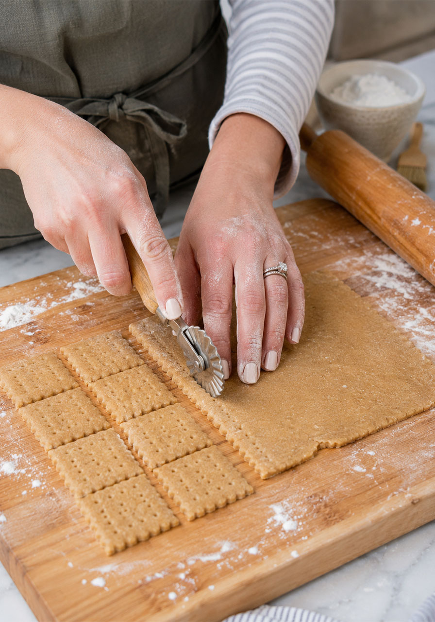 Hands cutting homemade honey graham cracker dough into squares with a pastry wheel on a floured board