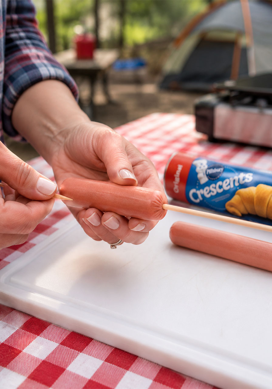 Hands inserting a wooden skewer three-quarters through a raw hot dog for campfire cooking