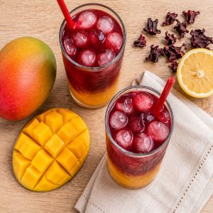 Overhead flat lay of two finished glasses of mango hibiscus iced tea with fresh mango, dried hibiscus flowers, and lemon on a wooden table