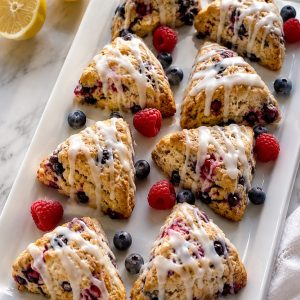 Mini mixed berry scones with lemon glaze arranged on a white party serving platter with fresh blueberries and raspberries