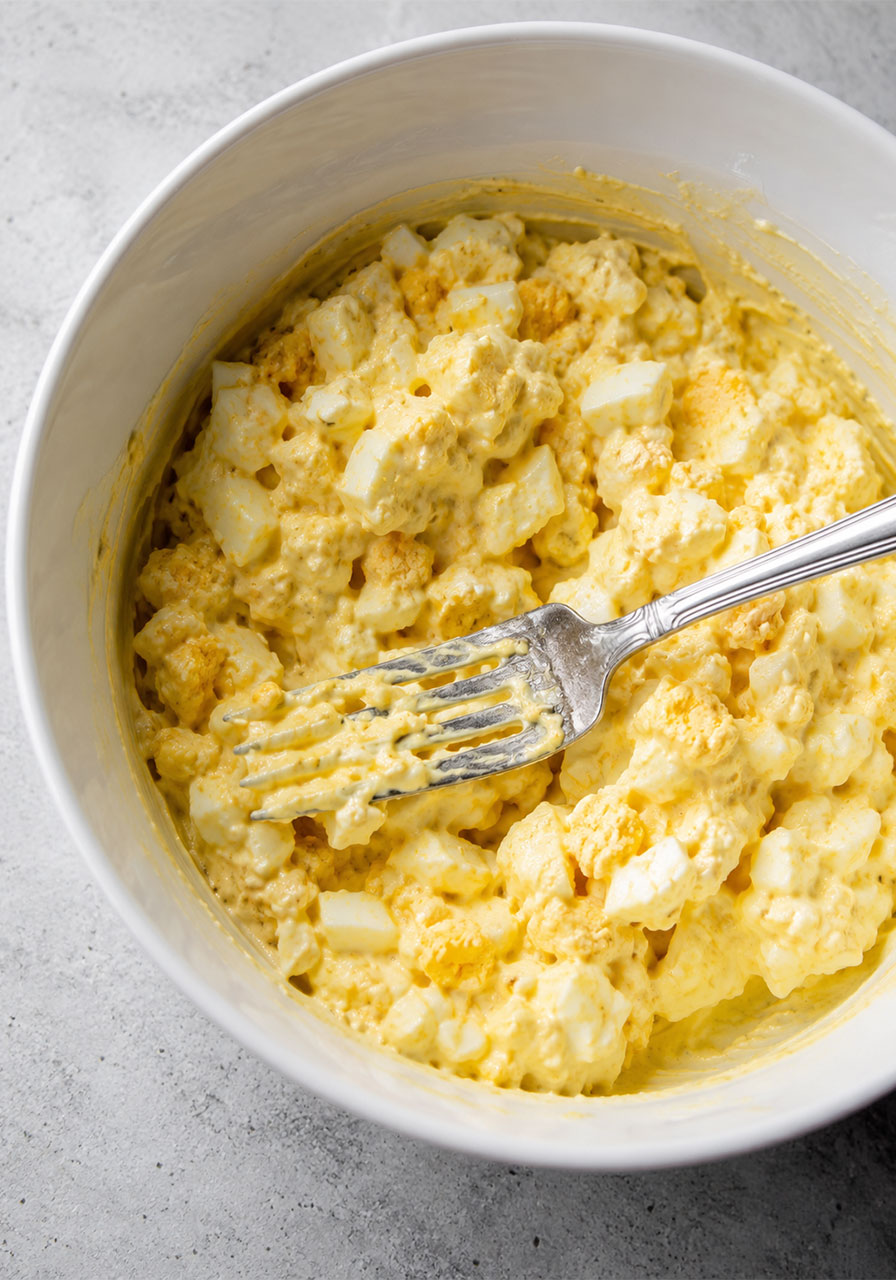Creamy egg salad mixture with mayo, Dijon mustard, and chopped hard boiled eggs in a white mixing bowl