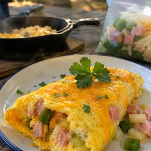 Finished omelet in a bag served on a white enamel camping plate outdoors, filled with ham, green pepper, and melted cheddar cheese