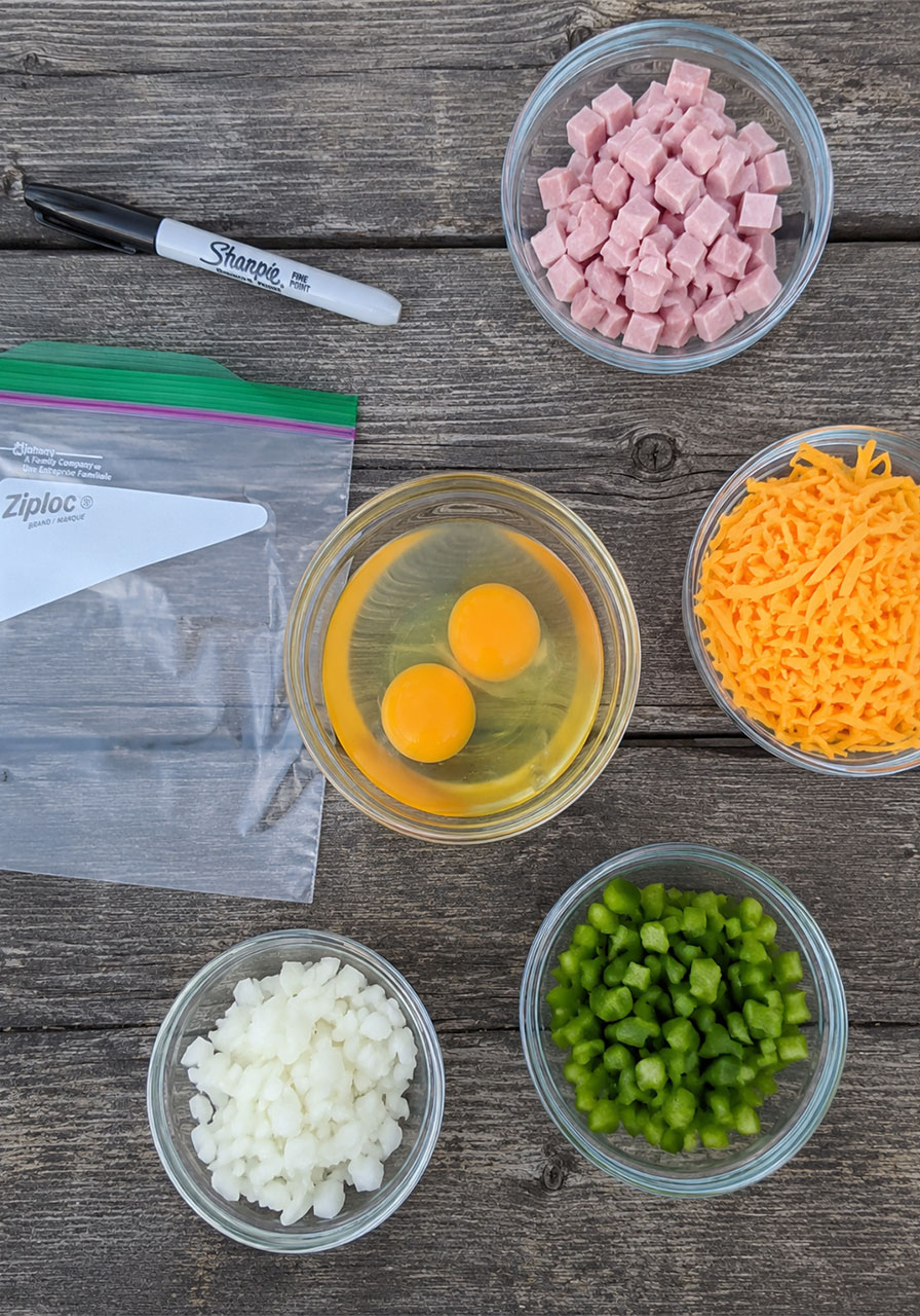 Flat lay of omelet in a bag ingredients on a picnic table including eggs, Ziploc bag, ham, cheddar cheese, green pepper, and onion