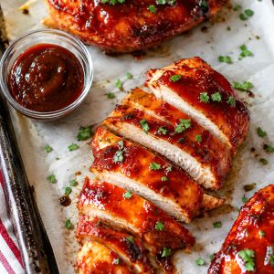 Juicy oven roasted BBQ chicken breasts sliced on a parchment-lined sheet pan with caramelized BBQ glaze and fresh parsley