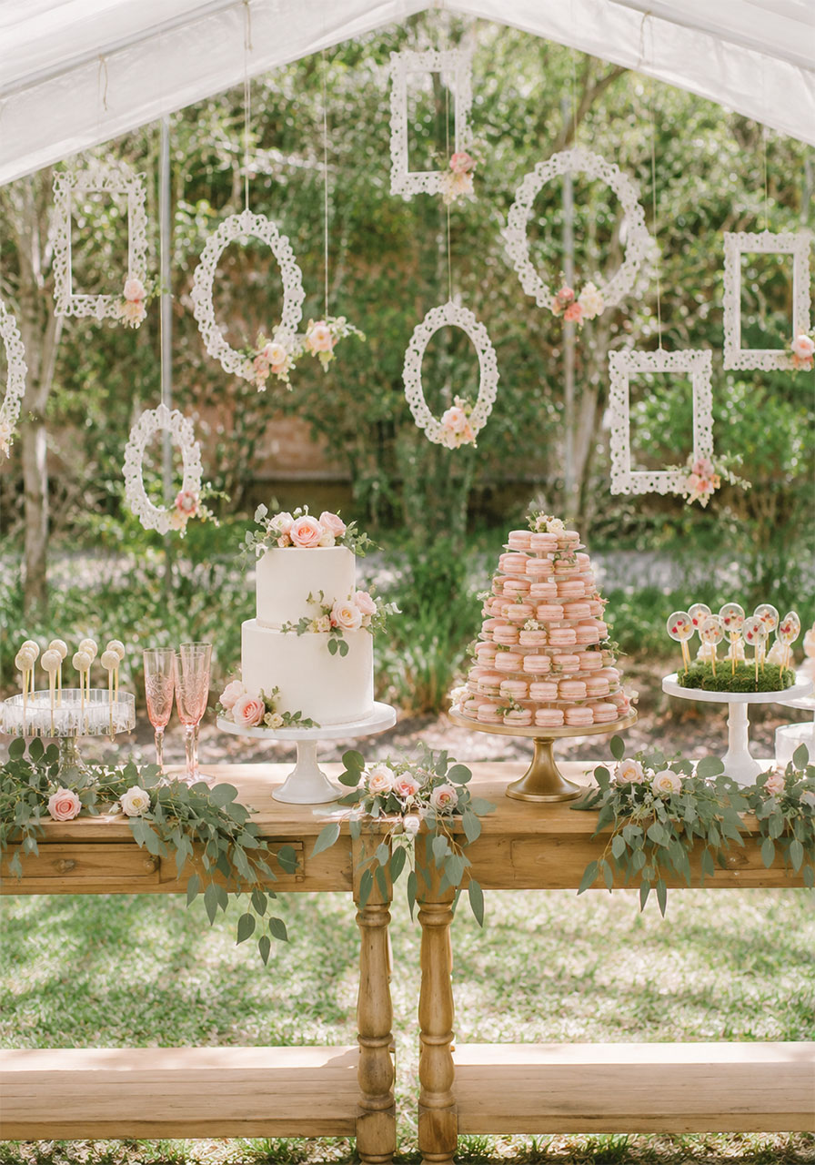 Romantic overgrown outdoor garden bridal shower dessert table with eucalyptus, macarons, and white cake under sheer canopy