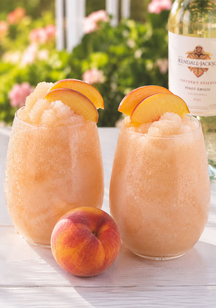 Two frosty blush peach wine slushies in stemless wine glasses garnished with fresh peach slices, on a bright outdoor table at a summer bachelorette party