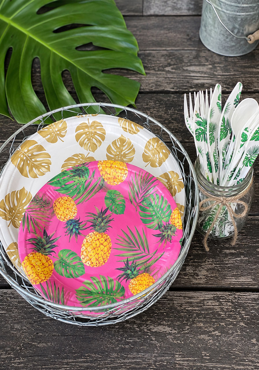 Pink pineapple-print party plates in wire basket with tropical leaf printed silverware in mason jar for birthday snack bar