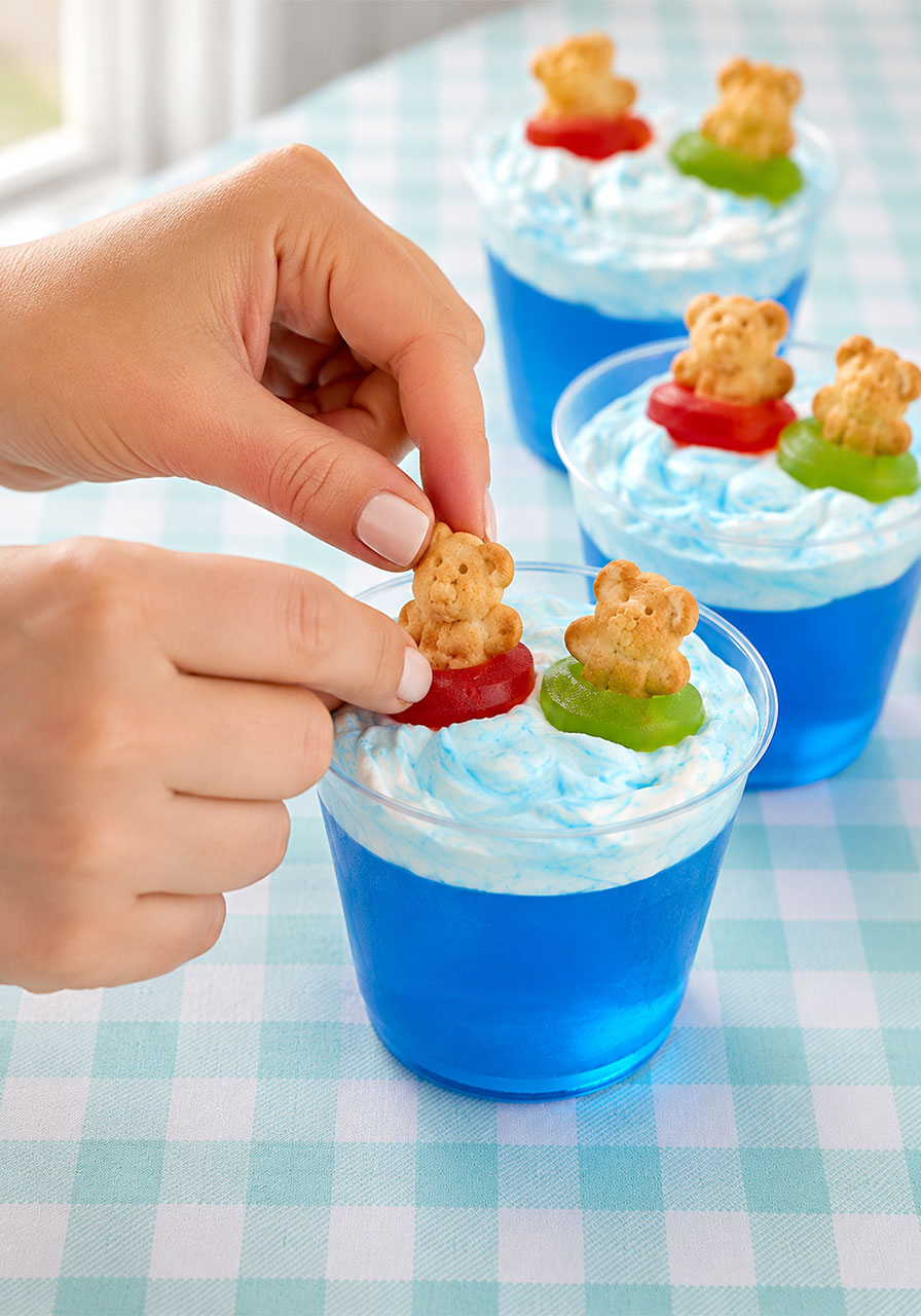 Woman's hands placing a Teddy Graham bear in a gummy Lifesaver floatie onto a blue Jello cup – final step of the cute pool party dessert recipe