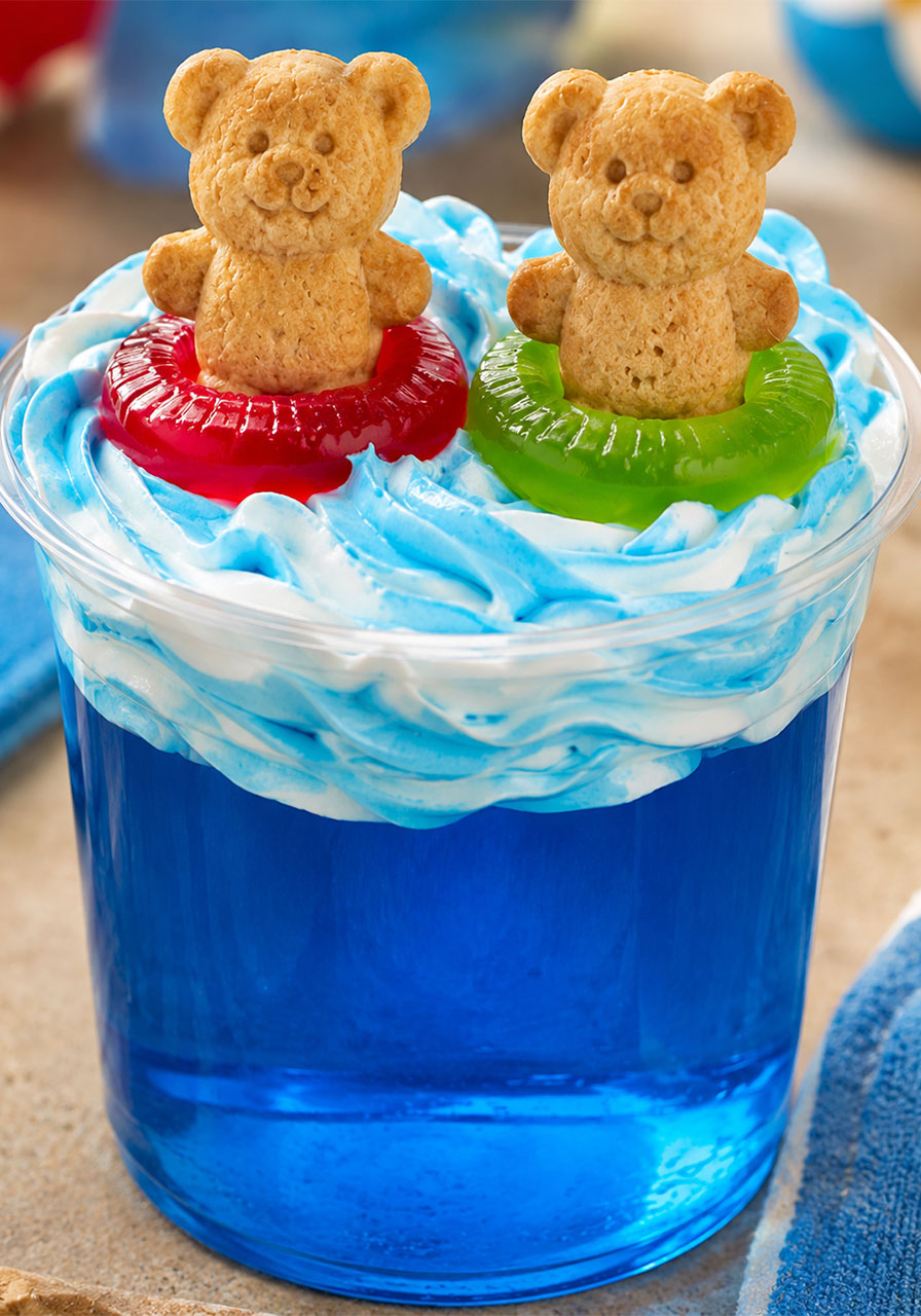 Close-up of a single pool party Jello cup with blue-swirled whipped topping waves and two Teddy Graham bears in gummy Lifesaver floaties – cute summer dessert idea