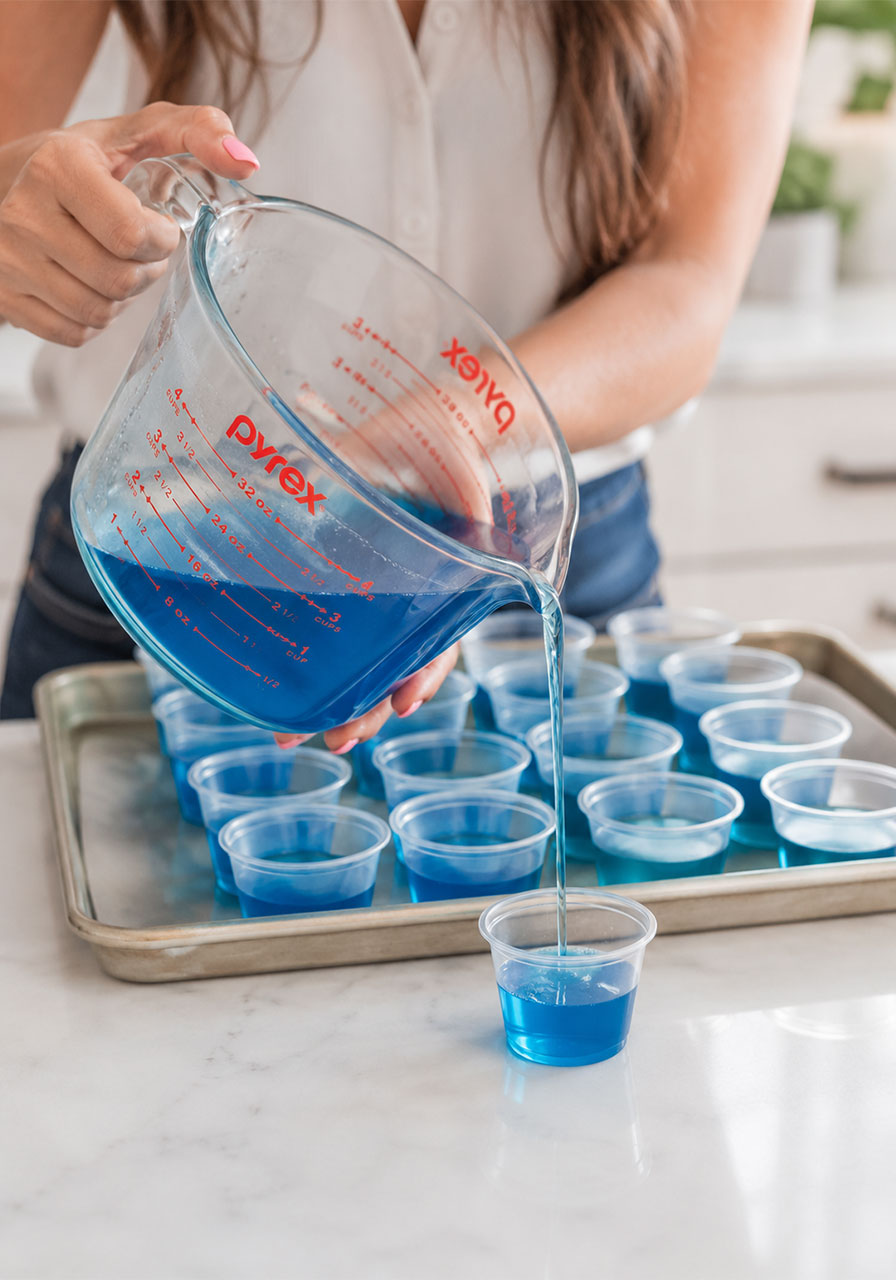 Pouring blue raspberry jello mixture into small plastic shot cups to make pool party jello shots