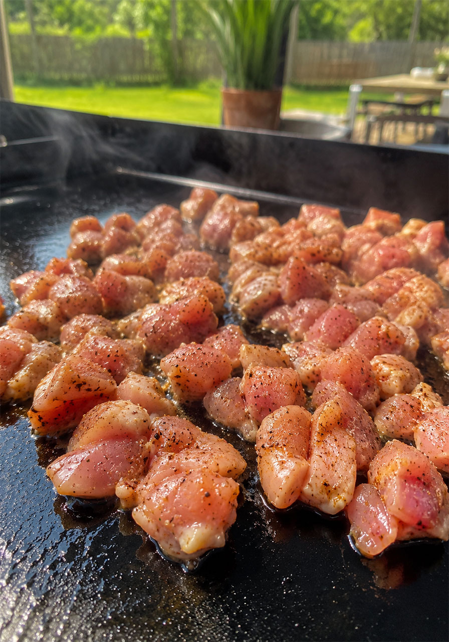 Raw diced chicken thighs seasoned with BBQ spice blend sizzling on a Blackstone flat top griddle