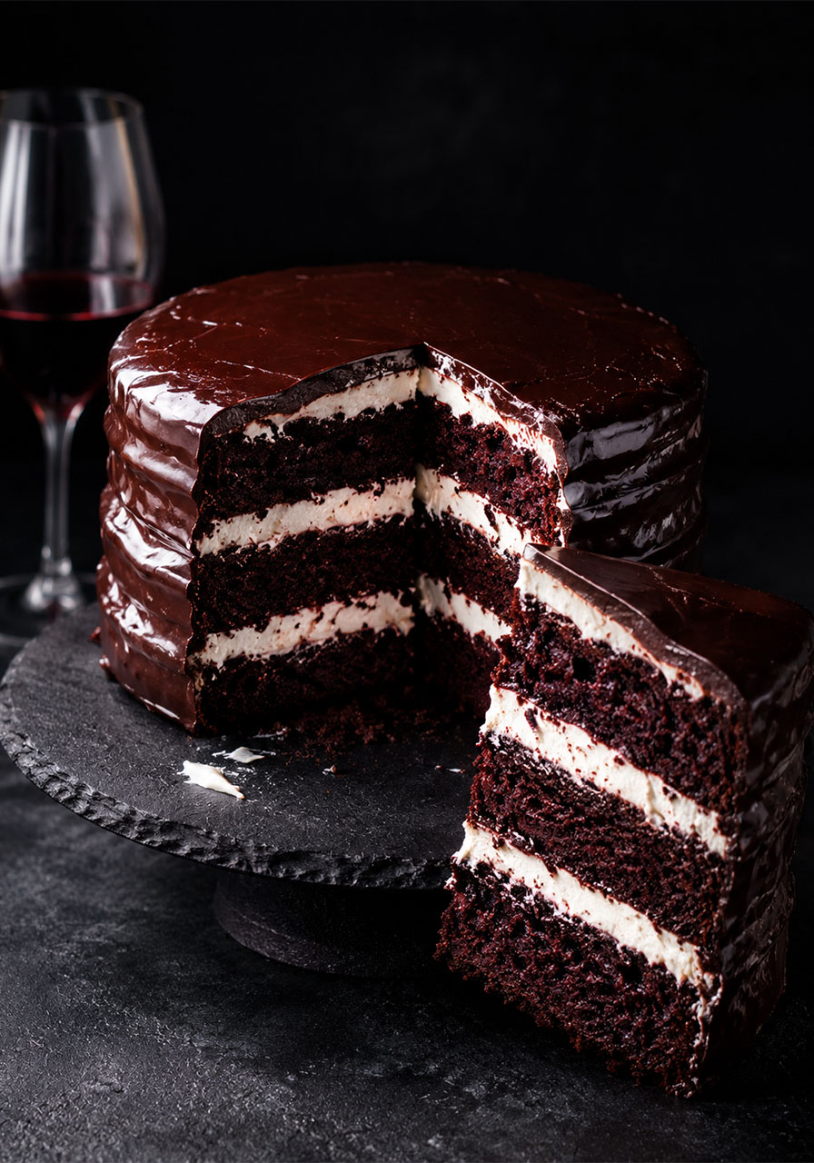 Three-layer Red Wine Velvet Cake with mascarpone filling, sliced open to show dark chocolate layers, on a slate stand with a glass of red wine in the background
