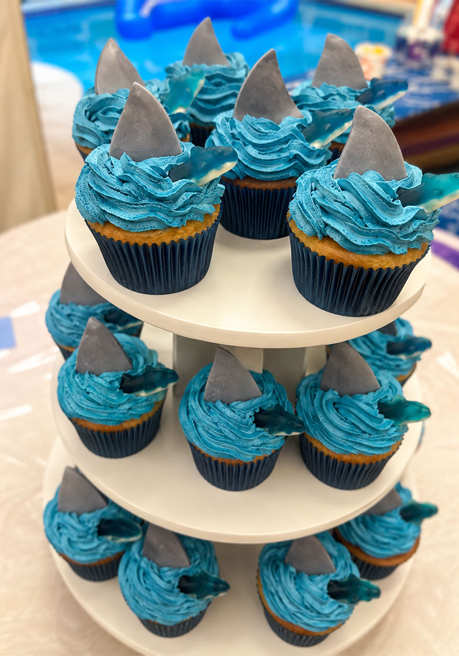 Shark cupcakes with blue wave buttercream frosting gray fondant fin and gummy shark on three tier stand for kids shark birthday party
