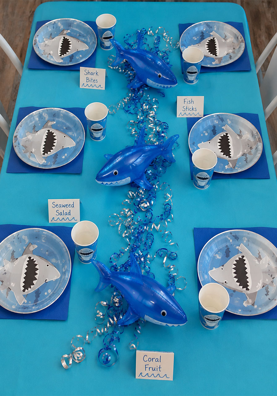 Shark themed kids birthday party table setting with shark print plates cups napkins teal tablecloth and inflatable shark centerpieces