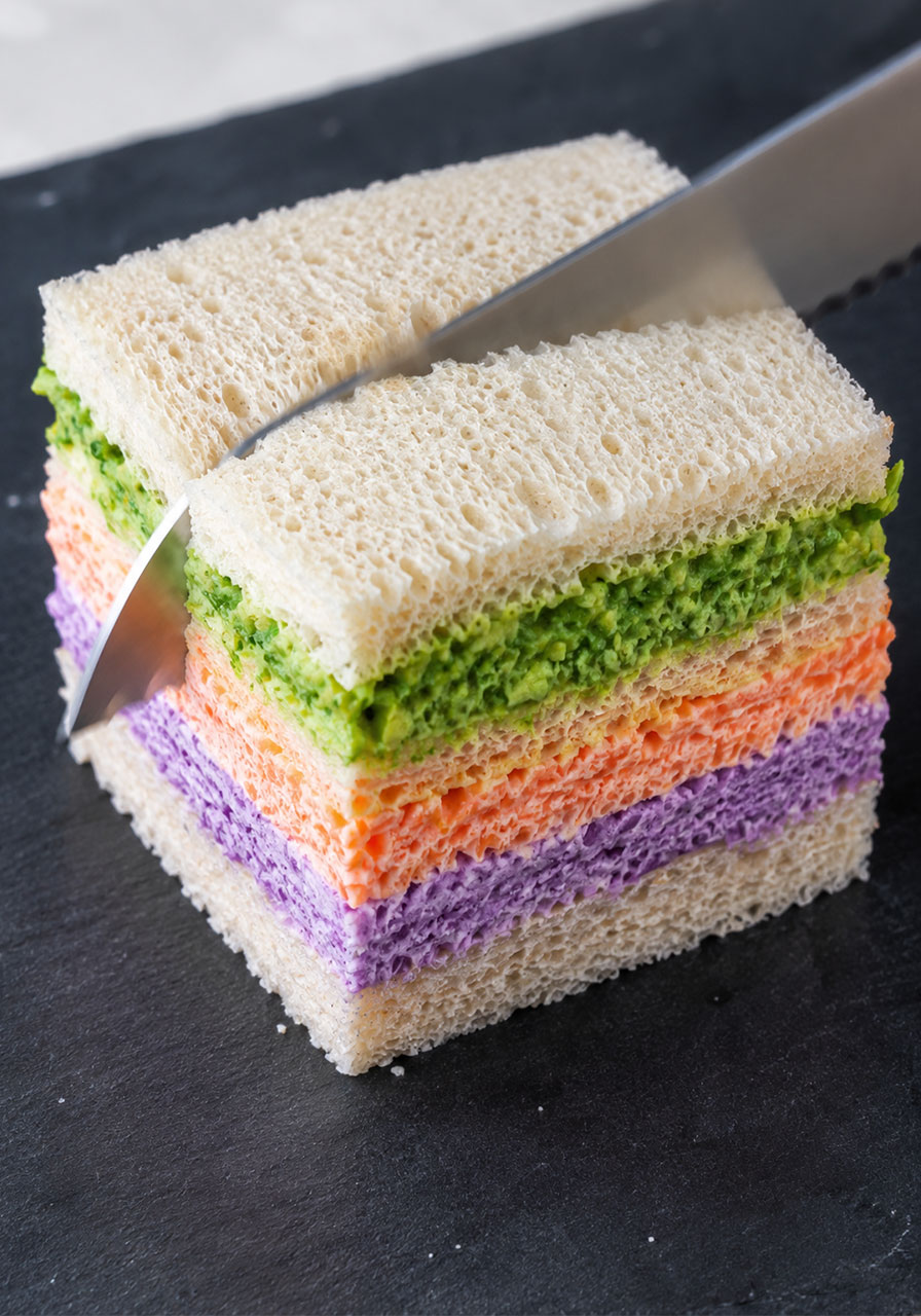 Serrated knife slicing through a stacked colorful tea sandwich to reveal green, orange, and purple yogurt layers inside